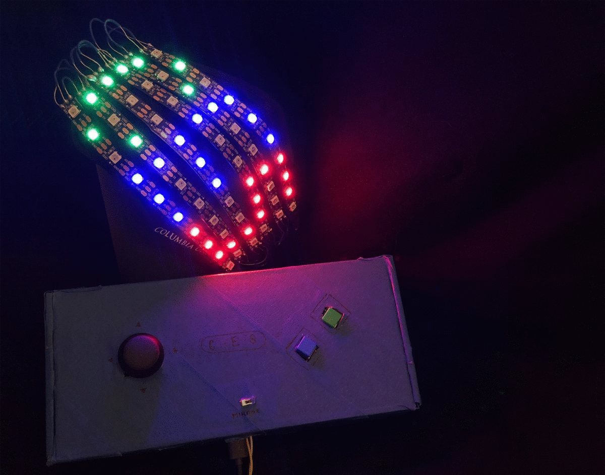 A picture of a COVID-19 facemask with strips of LEDs along the front. The picture is animated to show how the mask lights up and each of the individual LEDs can display its own color. The mask writes out the letters C E S, and below the mask is a blue rectangle with a joystick on the left and two square buttons on the right. This controller has a switch near the bottom edge and wires poking out of the bottom.